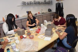ARTS 1 Students collaborate on pottery projects in a bright art classroom. Four young women work with clay, shaping pots and boxes. Supplies surround them on the table, with sinks and storage in the background.