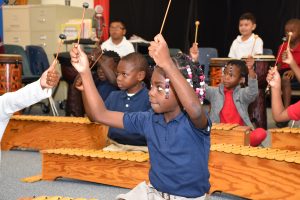 ARTS 2 A young Black girl plays a xylophone with enthusiasm in a music class, surrounded by other children holding mallets. The classroom features wooden xylophones and a file cabinet.