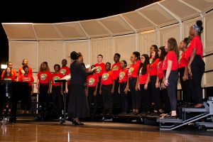 ARTS 3 A diverse youth choir, wearing matching red t-shirts, performs on a stage with a conductor leading them. A pianist is visible to the left.