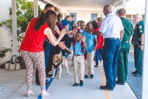 Mental Health 3 Elementary school students, including a young boy in a blue shirt, receive high fives from adults and a therapy dog as they enter the school. A line of uniformed officers stand in the background.