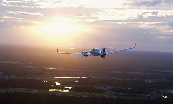 Prescott Diamond Diamond DA42 Twin Star soars above a lush landscape at sunset. The sun illuminates the aircraft, casting a warm glow over the trees and water below.