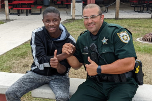 SAFE 2 A smiling Black teenager and a uniformed sheriff's deputy give thumbs up while sitting together outside. The deputy wears a green uniform with a badge. They clasp hands in a gesture of camaraderie.