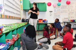 TEACH 2 A teacher instructs a diverse group of elementary students on the floor in a colorful classroom. Math concepts are visible on the whiteboard, and learning materials are organized in colorful bins.