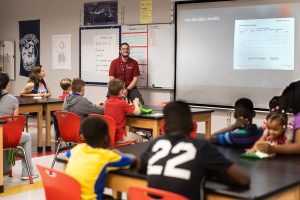 TEACH 3 A diverse group of middle school students sit at desks in a classroom, listening to a male teacher present a "Decision Matrix" on a projector screen. The teacher wears a red polo shirt.