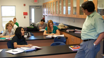Classroom A high school science class in session: students at desks listen to their teacher lecturing in a bright classroom, with lab counters and cabinets visible.