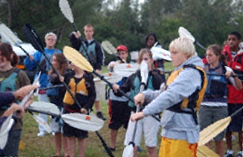 Paddle Group of kids outdoors holding kayak paddles, practicing strokes on land before getting in the water, wearing life vests, learning kayaking techniques.