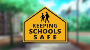 School Safety Yellow road sign with a black border reads "Keeping Schools Safe" above a silhouette of an adult and child walking. A blurred school building and trees are in the background.