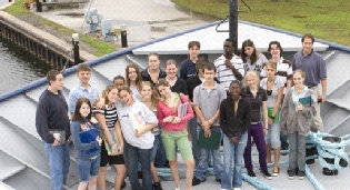 Ship A diverse group of high school students and two adults pose on the deck of a canal boat. They are smiling and holding notebooks, with a canal lock visible behind them.