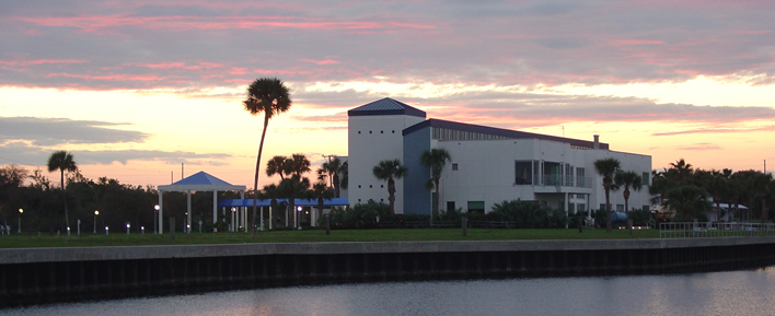 Sunset Waterfront building at sunset with palm trees, blue accents, and a covered walkway. Pink and orange hues fill the sky, reflecting on the water.